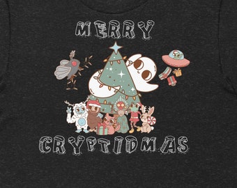 May include: A dark heather grey t-shirt with a white graphic of a Christmas tree with a ghost, a moth, a yeti, a sasquatch, a chupacabra, a jackalope, and a Bigfoot. The text "Merry Cryptidsmas" is printed below the graphic.