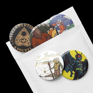 May include: Four round button pins with different designs. The first pin is black with gold text that reads "Cryptid Apparel" and a gold illustration of a crescent moon, eye, and a triangle with a star inside. The second pin is a white background with a brown and green illustration of a frog holding a stick. The text reads "The Loveland Frogman". The third pin is a yellow background with a black illustration of a creature with wings and red eyes. The fourth pin is a brown background with a brown and orange illustration of a creature with brown fur and a white beard.
