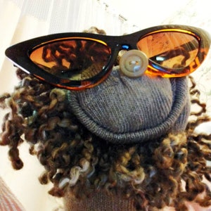 May include: A brown and tan sock puppet wearing a pair of brown cat-eye sunglasses with orange lenses. The puppet has a button nose and a fuzzy brown beard.