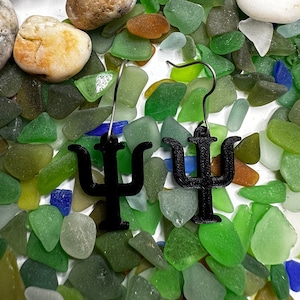 Puede incluir: Un par de pendientes de metal negro con forma de la letra griega Psi, sobre un fondo de vidrio de mar verde, azul y blanco.