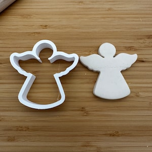 Puede incluir: Un cortador de galletas de ángel de plástico blanco y un recorte de ángel de arcilla blanca sobre una superficie de madera.