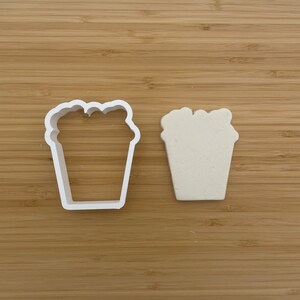 May include: A white plastic cookie cutter shaped like a bucket of popcorn. The cutter is shown next to a white plastic cookie that has been cut out using the cutter.