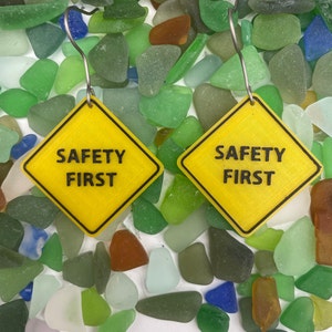 May include: A pair of yellow safety sign earrings with the text "SAFETY FIRST" on each sign. The earrings are displayed on a bed of colorful sea glass.