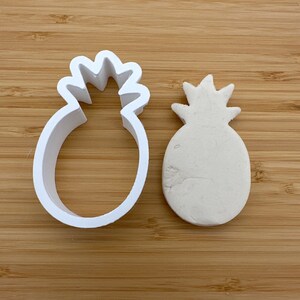 May include: White plastic pineapple cookie cutter and a white molded pineapple shape on a wooden surface.