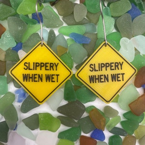 Puede incluir: Dos pendientes amarillos en forma de diamante con el texto "SLIPPERY WHEN WET" en letras negras. Los pendientes cuelgan de ganchos plateados y están rodeados de vidrio de mar.