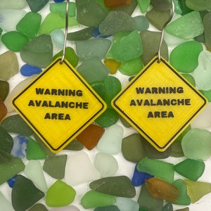 May include: A pair of yellow warning sign earrings with the text "WARNING AVALANCHE AREA" on each earring. The earrings are displayed on a bed of green, blue, and brown sea glass.