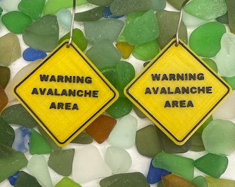 Puede incluir: Un par de pendientes de señal de advertencia amarilla con el texto "WARNING AVALANCHE AREA" en cada pendiente. Los pendientes se muestran sobre un lecho de vidrio de mar verde, azul y marrón.