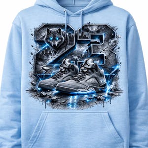 以下が含まれることがあります： オオカミ、数字の23、スニーカーをあしらったグラフィックデザインのライトブルーのパーカー。デザインには都市景観、稲妻、ひび割れた地球の要素が含まれています。スニーカーには数字の23が入っています。