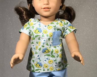 Puede incluir: Una muñeca con un top de uniforme médico de estampado floral verde y azul claro con un bolsillo azul, pantalones azules claros y zapatillas blancas.