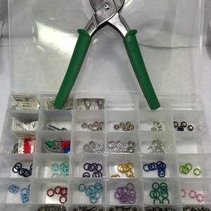 May include: A clear plastic organizer filled with various snap fasteners and tools. The organizer contains a variety of metal snaps, colored rings, and a green-handled snap pliers tool. The snaps are in silver, gold, blue, green, and purple.