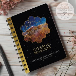 Op de afbeelding: Een zwart spiraalnotitieblok met een gouden rand. De omslag heeft een kleurrijk beeld van de kosmos met een honingraatpatroon eroverheen. De tekst "COSMIC REFLECTIONS JAMES WEBB SPACE TELESCOPE EST. JULY 12, 2022" is in goud op de omslag gedrukt.