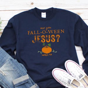 May include: Navy blue sweatshirt with the text "are you FALL-O-WEEN JESUS?" and a pumpkin graphic. The sweatshirt is paired with blue jeans and white sneakers, creating a casual fall outfit.
