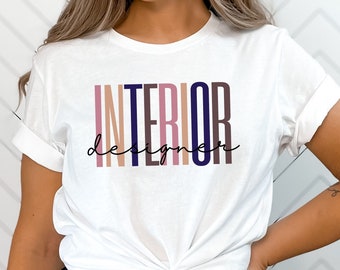 Könnte beinhalten: Weißes T-Shirt mit dem Text "INTERIOR DESIGNER" in einem mehrfarbigen Design. Das Wort "INTERIOR" steht in vertikalen Blockbuchstaben, "DESIGNER" in Schreibschrift. Das Shirt ist an der Taille geknotet.