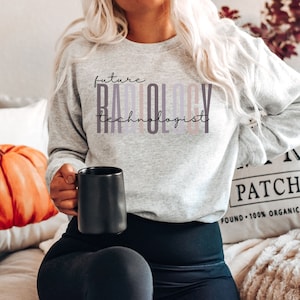 May include: A gray sweatshirt with the text "future radiology technologist" printed on it in a colorful, layered font. The sweatshirt is being worn by a person holding a black mug.