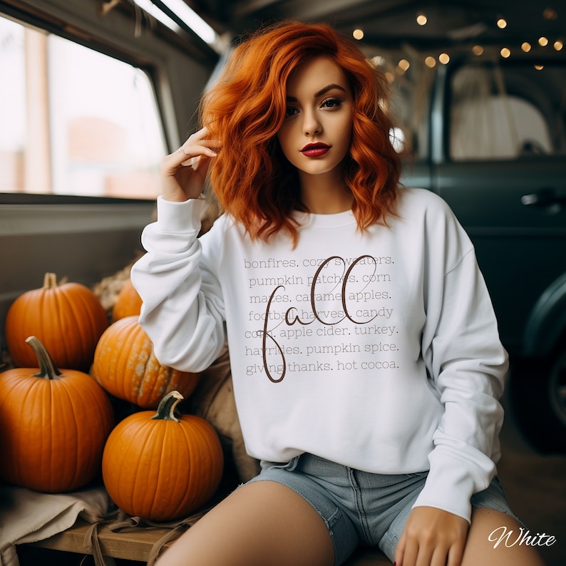 May include: A white sweatshirt with the word "Fall" in a cursive font, listing autumn-themed words. The sweatshirt is worn with denim shorts. Several orange pumpkins are arranged on a wooden surface.