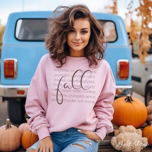 May include: A light pink sweatshirt with the word "Fall" in a cursive font, surrounded by a list of autumn-themed words. The model is wearing blue jeans and is sitting in front of a blue van and pumpkins.
