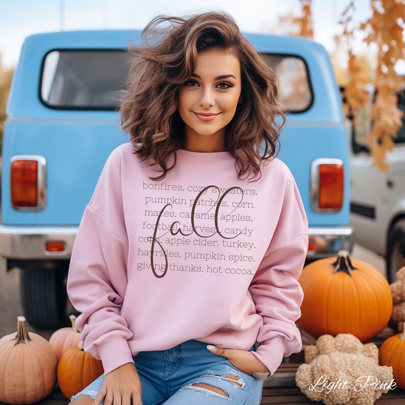 May include: A light pink sweatshirt with the word "Fall" in a cursive font, surrounded by a list of autumn-themed words. The model is wearing blue jeans and is sitting in front of a blue van and pumpkins.