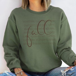 May include: An olive green sweatshirt with the word "fall" in a cursive font. The sweatshirt has a list of fall-themed words in a darker brown font, including "bonfires," "pumpkin patches," and "apple cider."