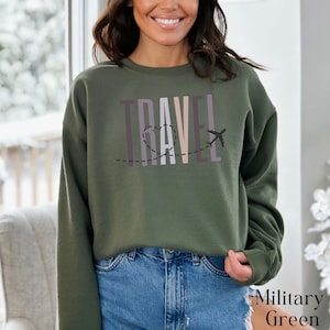 May include: A green sweatshirt with the word "TRAVEL" printed in colorful letters with a plane and a heart design. The sweatshirt is being modeled by a person wearing blue jeans.
