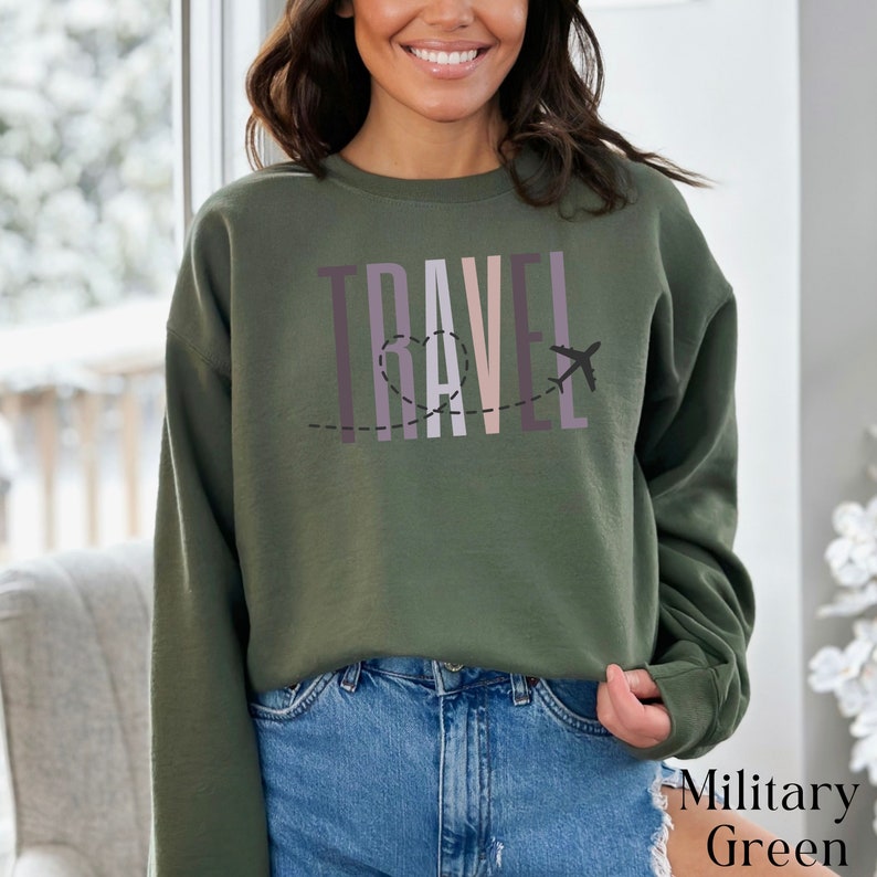 May include: A green sweatshirt with the word "TRAVEL" printed in colorful letters with a plane and a heart design. The sweatshirt is being modeled by a person wearing blue jeans.