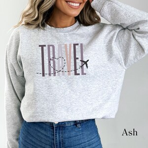 May include: A gray sweatshirt with the word "TRAVEL" printed in colorful letters. A small airplane and a heart are drawn above the word. The sweatshirt is being worn by a person.