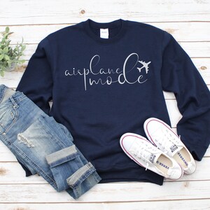 May include: Navy blue sweatshirt featuring the text "airplane mode" in a cursive script, accompanied by a small aeroplane graphic. The image includes blue jeans and white trainers on a light wood surface.