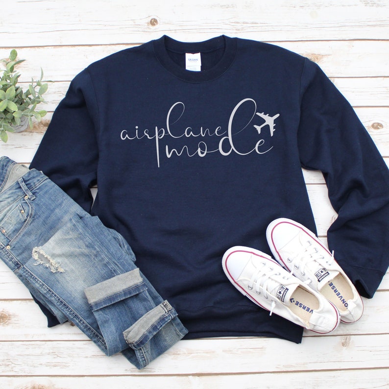 May include: Navy blue sweatshirt featuring the text "airplane mode" in a cursive script, accompanied by a small aeroplane graphic. The image includes blue jeans and white trainers on a light wood surface.
