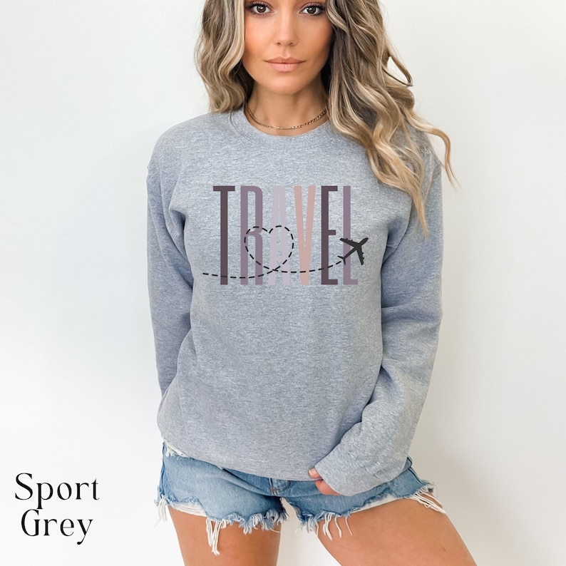 May include: A heather grey sweatshirt with the word "TRAVEL" printed in colorful vertical stripes. A small airplane with a dotted line trail forms a heart shape above the word.