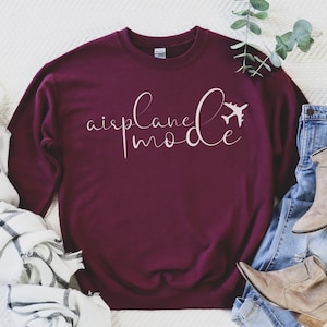 May include: A burgundy sweatshirt featuring the text "airplane mode" in a cursive script, with an aeroplane icon. The garment is shown with jeans and boots on a white background.