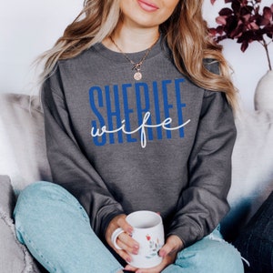 May include: A gray sweatshirt with the text "SHERIFF wife" in blue and white lettering.
