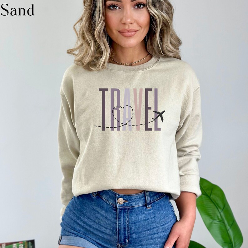 May include: A beige sweatshirt with a graphic print of the word "TRAVEL" in colorful letters with a heart and airplane design.