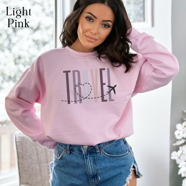 May include: A light pink sweatshirt with the word "TRAVEL" printed on the front in a colorful, layered font. A small airplane and heart are included in the design.
