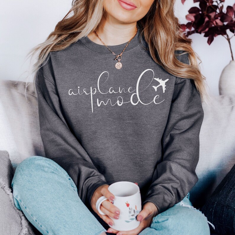 May include: A charcoal grey sweatshirt featuring the words "airplane mode" in white script, with a small airplane graphic. The long-sleeved sweatshirt has a crew neck. The person is holding a white mug with a floral design.
