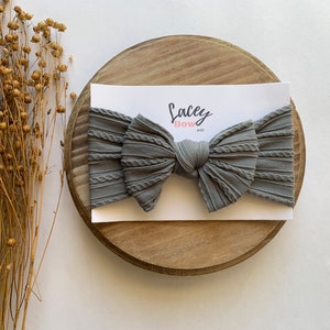 May include: A grey, textured, knotted hair bow with the text "Lacey Bow Co." on a white card.