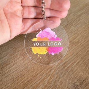 May include: Clear acrylic keychain with a silver chain. The keychain features a colorful design with pink, yellow, and orange paint splatters and the text "YOUR LOGO". A perfect accessory for keys or bags.