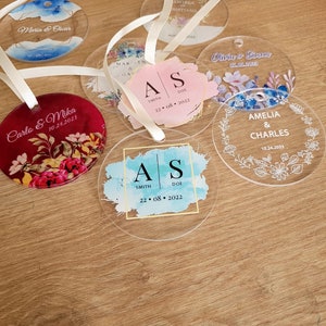 May include: Six clear acrylic wedding favour ornaments with floral designs and the names of the couples and their wedding dates. The ornaments are decorated with watercolour-style backgrounds and gold accents.