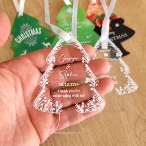 Può includere: Un ornamento per albero di Natale in acrilico trasparente con un design bianco opaco. L'ornamento presenta i nomi "George & Sophia" e la data "20.12.2024". È incluso anche il testo "Grazie per aver festeggiato con noi!"