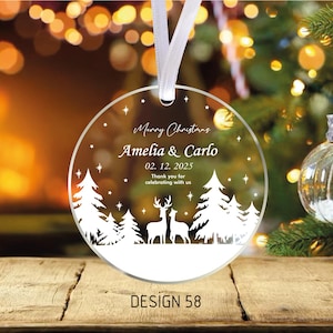 May include: A clear, round Christmas ornament with a white ribbon. The ornament features a winter scene with trees, deer, and the text "Merry Christmas Amelia & Carlo 02.12.2025". The design is labeled "DESIGN 58".