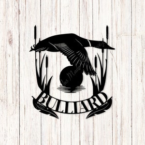 Puede incluir: Silueta negra de metal de un pato volando sobre un estanque con juncos y la palabra "BULLIARD" en una pancarta debajo.