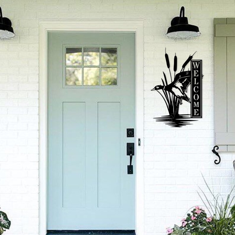 May include: Black metal welcome sign depicting a duck in flight, reeds, and the word "WELCOME" in a vertical format. The sign is mounted on a white brick wall next to a pale blue door.