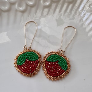 May include: A pair of strawberry earrings. Each earring has a red beaded strawberry design with green beaded leaves and a gold beaded border. The earrings have gold-coloured ear wires.