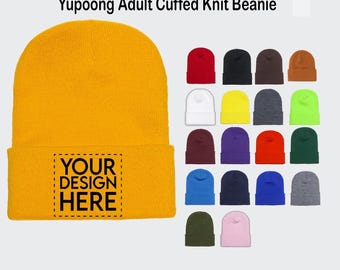 May include: A yellow knit beanie with a fold-over cuff. The beanie has a blank space for customization with the text "YOUR DESIGN HERE". The image also shows a variety of colors for the beanie.