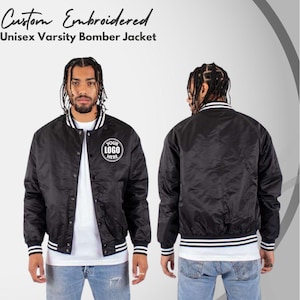 May include: A black varsity bomber jacket with white striped ribbed cuffs and collar. The front features a circular patch with the text "YOUR LOGO HERE". The jacket is shown from both the front and back views, displaying a snap button closure.