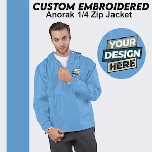 May include: A light blue 1/4 zip jacket with a hood. The jacket has a zipper closure and a pocket on the left chest. The text "YOUR DESIGN HERE" is embroidered on the pocket.