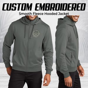 Pode incluir: Casaco cinzento com capuz em fleece liso com fecho de correr completo. O casaco apresenta um emblema bordado no peito esquerdo e o texto "CUSTOM EMBROIDERED" acima de "Smooth Fleece Hooded Jacket".