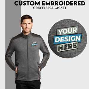May include: A gray fleece jacket with a zipper closure and two pockets. The jacket has a contrasting black panel on the shoulders and sleeves. The text "YOUR DESIGN HERE" is embroidered on the jacket in blue and white.