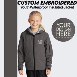May include: A gray youth waterproof insulated jacket with a zipper closure and a hood. The jacket has two pockets on the front and one pocket on the left sleeve. The text "YOUR DESIGN HERE" is embroidered on the front of the jacket.