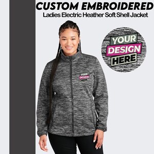 May include: A black and gray soft shell jacket with a zipper closure. The jacket has a mock neck and two front pockets. The jacket is embroidered with the text "YOUR DESIGN HERE" in pink and black.