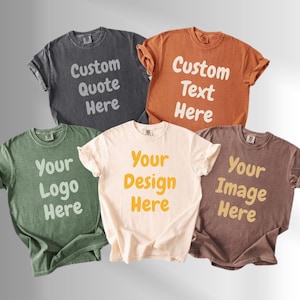 May include: Five short-sleeve crew neck t-shirts in charcoal, orange, green, cream, and brown. Each shirt features bold, rounded white or yellow text printed on the chest, including phrases like Custom Quote Here, Custom Text Here, and Your Design Here.