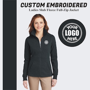 May include: A dark gray fleece full-zip jacket with a mock neck collar. The jacket has two front pockets and a zipper closure. The jacket has a white embroidered logo on the left chest that says "YOUR LOGO HERE".